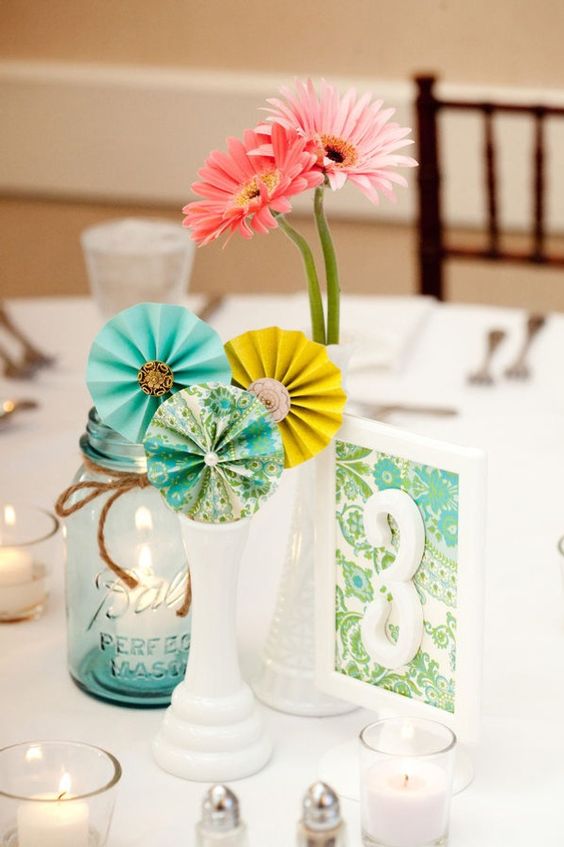a simple and pretty wedding centerpiece of a vase with turquoise and yellow paper fans, with pink blooms and a candle in a jar