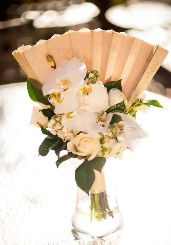 a simple and pretty wedding centerpiece with white roses and orchids, with a paper fan and leaves is a lovely solution