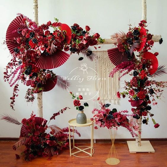 a super bold and catchy wedding arch decorated with deep red, black and burgundy blooms, with red paper fans and macrame, with gold furniture