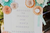 a wedding sign decorated with peachy and turquoise paper fans looks cute and cool and will make the space bolder