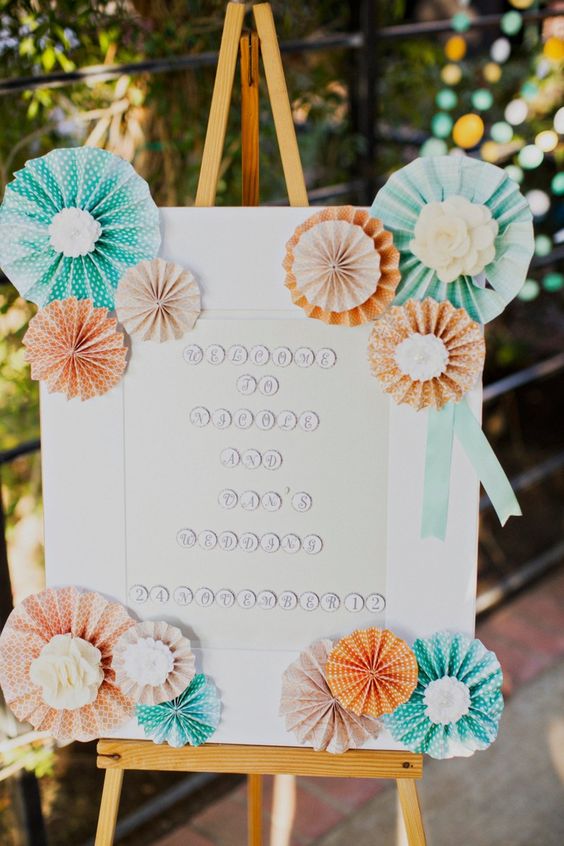 a wedding sign decorated with peachy and turquoise paper fans looks cute and cool and will make the space bolder