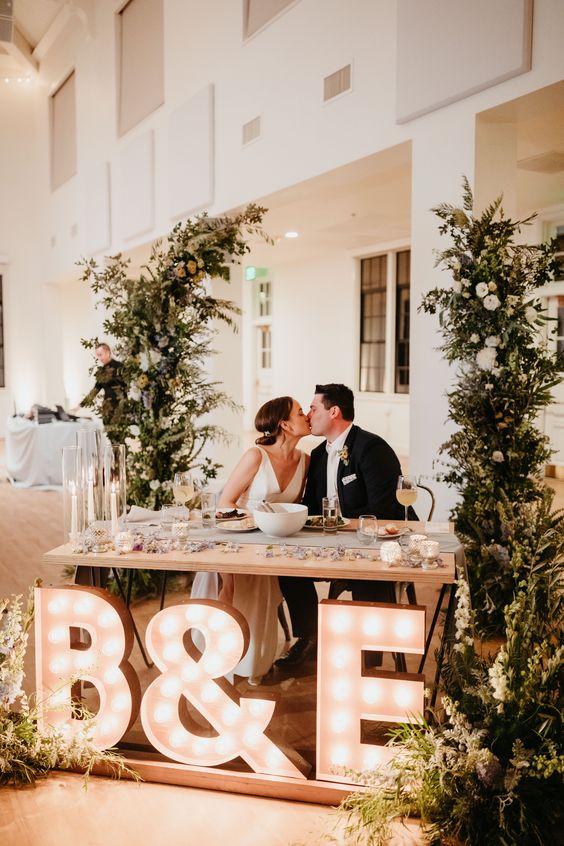 a wedding sweetheart table with greenery and neutral blooms around, plus oversized marquee monograms and an ampersand