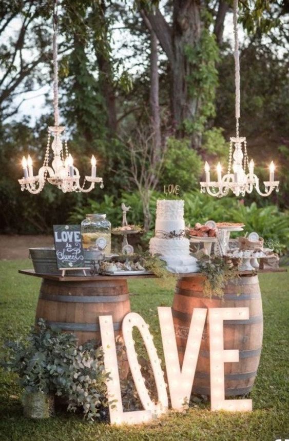 an outdoor rustic wedding dessert table placed on barrels, with greenery, marquee letters and chandeliers over it