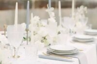 a beautiful and refined modern wedding tablescape with a white tablecloth and grey napkins, tall and thin candles, neutral blooms