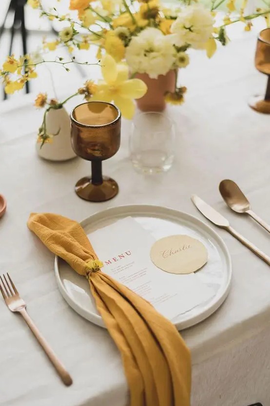 a beautiful spring wedding tablescape with a neutral tablecloth, a mustard napkin, a yellow and mustard centerpiece and a brown glass