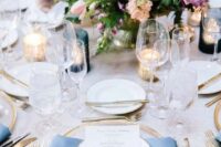 a bright and chic spring wedding tablescape with a pink tablecloth and blue napkins, bold blooms and greenery, candles and gold rimmed plates