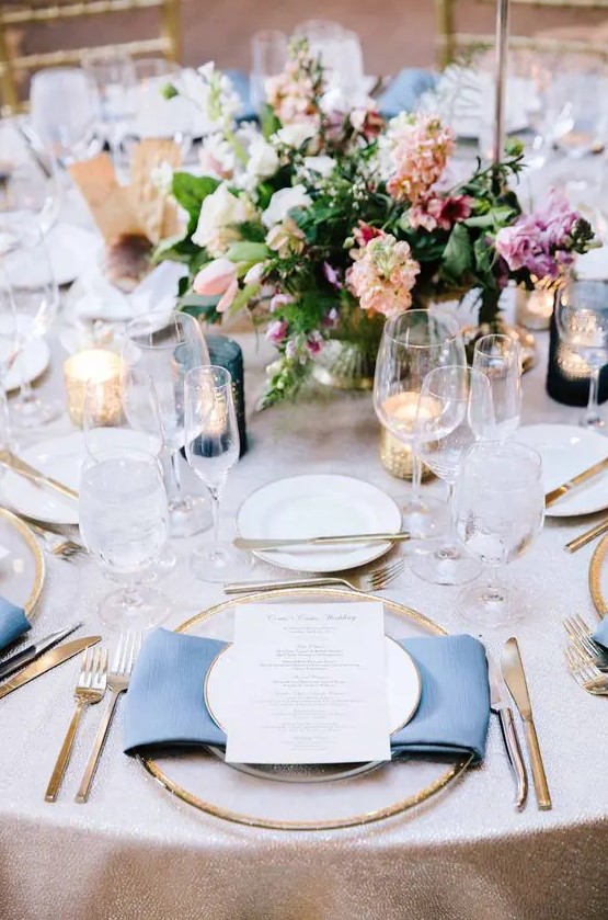 a bright and chic spring wedding tablescape with a pink tablecloth and blue napkins, bold blooms and greenery, candles and gold rimmed plates