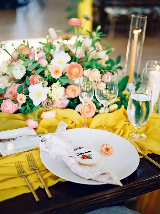a bright wedding table setting with a yellow table runner, pink and red blooms, gold cutlery and a cookie