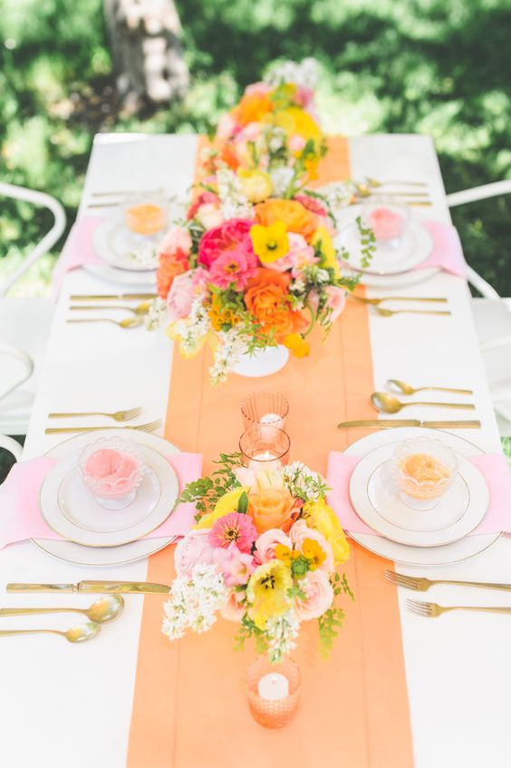 a bright wedding tablescape with an orange runner, pink napkins, pink, yellow and orange blooms and candles in peachy candleholders