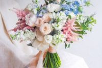 a chic pastel spring wedding bouquet with peachy, pink, blue and white blooms and greenery plus long ribbons