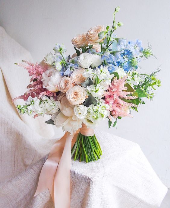 a chic pastel spring wedding bouquet with peachy, pink, blue and white blooms and greenery plus long ribbons