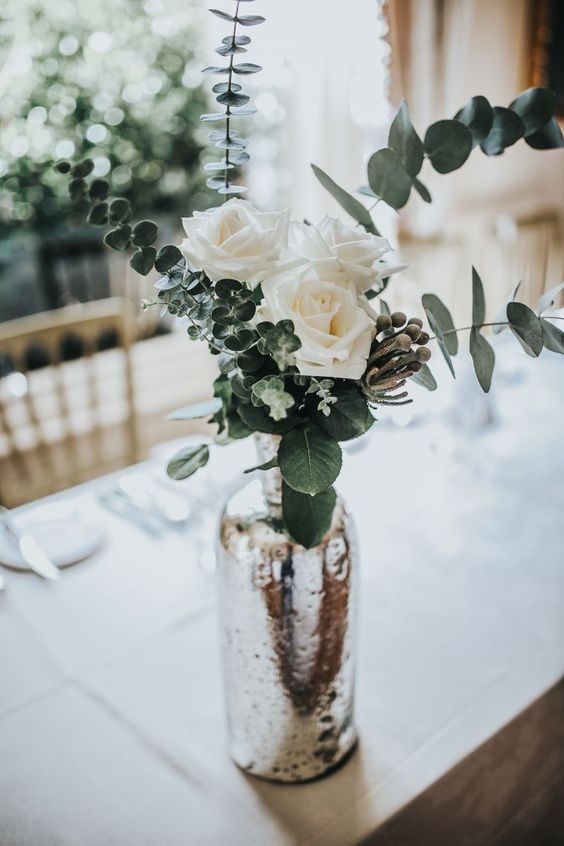 a chic wedding centerpiece of a mercury glass vase, greenery, berries, white roses is a very elegant and simple piece you can recreate