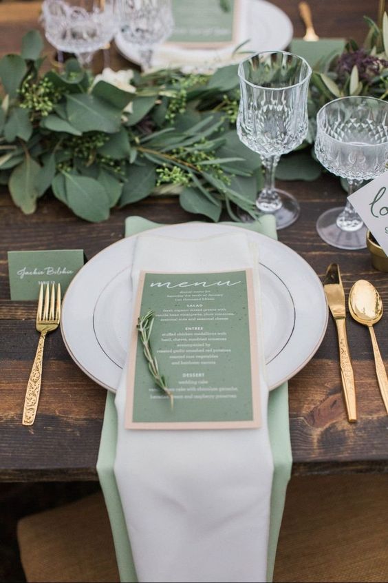 a chic wedding tablescape with a greenery runner, a sage green napkin, card and menu, elegant glasses and gold cutlery