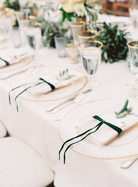 a chic wedding tablescape with creamy linens and green ribbons, a greeneyr runner and white blooms plus gold-rimmed glasses