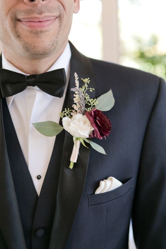 a classic navy tux with black lapels, a white and burgundy ranunculus, greenery and grasses are a chic and stylish combo to rock