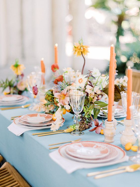 a colorful spring wedding tablescape with a blush tablecloth, peachy plates and white napkins, orange candles, neutral and bright blooms