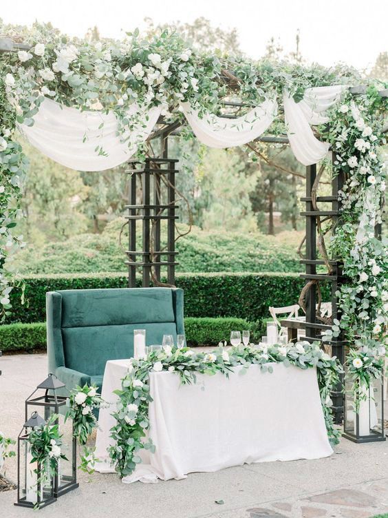 a gorgeous sweetheart table space with greenery, white blooms, an emerald sofa, a white table and candle lanterns