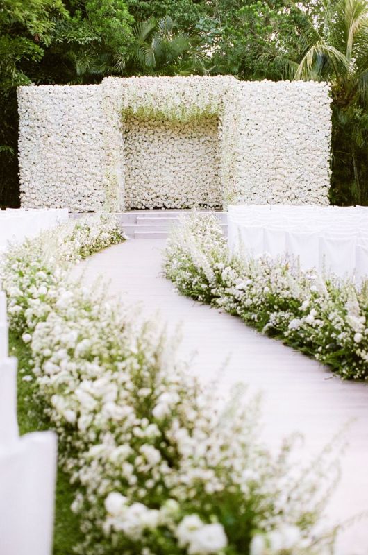 a luxurious wedding ceremony space covered with a white flower wall and greenery and white blooms lining up the aisle