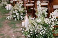 a natural wedding aisle with greenery and white blooms, with pillar candles is a very chic and cool idea for a spring or summer wedding