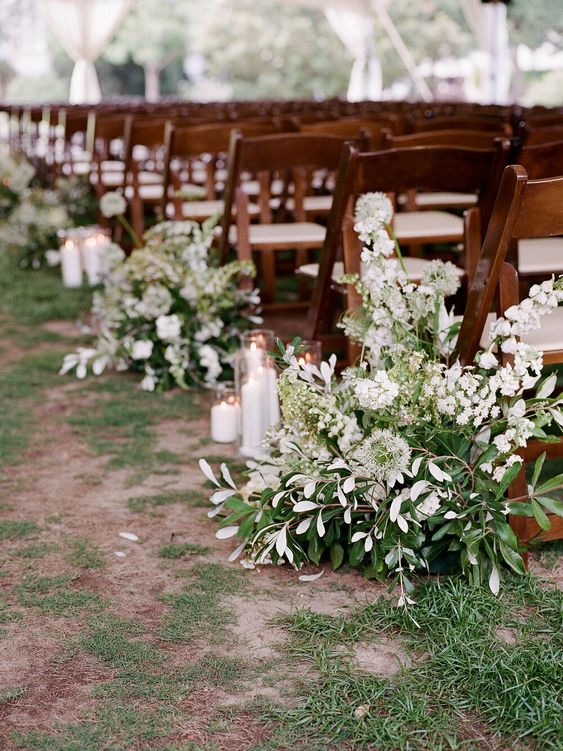 a natural wedding aisle with greenery and white blooms, with pillar candles is a very chic and cool idea for a spring or summer wedding