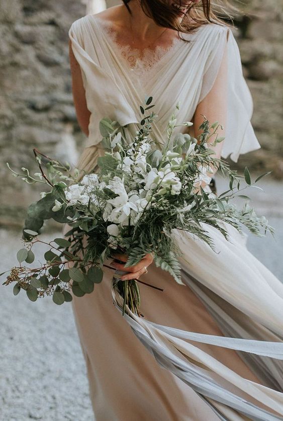 a neutral and airy wedding dress with draperies, with a greenery and white bloom wedding bouquet and grey ribbons
