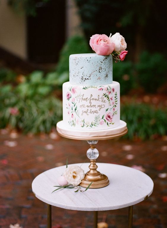 a pastel wedding cake with a dusty blue and gold leaf tier, with hand painted blooms and a quote plus some fresh blooms on top for a spring or summer wedding