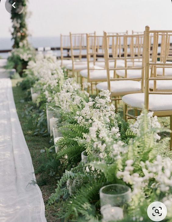 a pretty textural wedding aisle with white blooms and ferns plus pillar candles in glasses is a very elegant idea