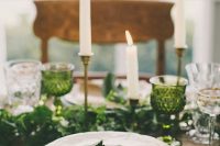 a pretty wedding table setting with a greenery runner and green glasses, neutral porcelain and candles and gold cutlery