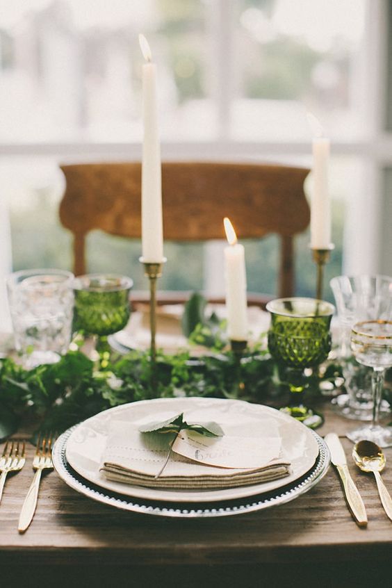 a pretty wedding table setting with a greenery runner and green glasses, neutral porcelain and candles and gold cutlery