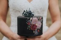 a small and delicate black wedding cake with painted pink and burgundy blooms gold leaf and some berries and thistles on top
