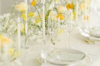 a spring wedding tablescape with yellow and white blooms, white plates and tlal and thin candles is very chic