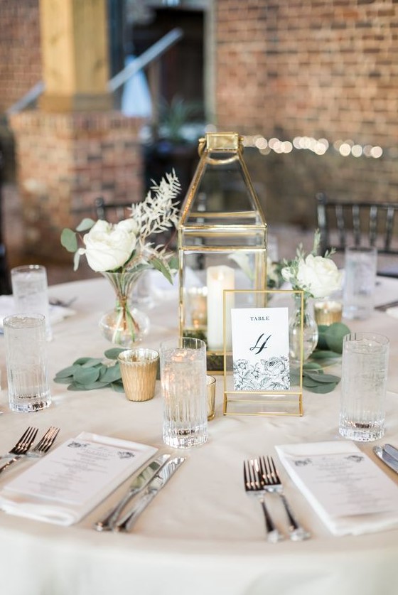 a stylish neutral wedding table with a gold candle lantern, a table number, white blooms and greenery and white linens