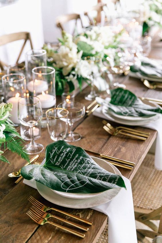 a stylish tropical wedding tablescape with an uncovered table, white porcelain, linens and candles, large leaves and white blooms and greenery