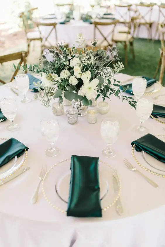 a stylish wedding table setting in white accented with emerald napkins and with a green and white wedding centerpiece with thistles