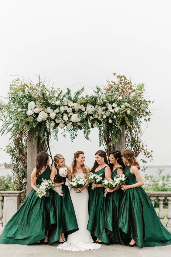 a wedding arch covered with lush greenery and white hydrangeas, a bride in white and bridesmaids is beautiful hunter green A-line high low dresses