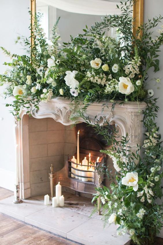 a wedding fireplace with various candles decorated with greenery and white blooms looking so natural as if they are growing here