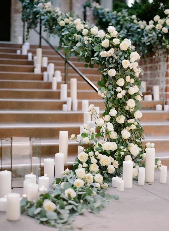 a wedding staircase decorated with greenery and white roses plus pillar candles and candle lanterns is a very chic idea