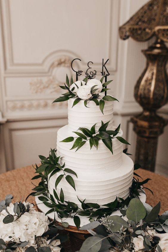 a white textural buttercream wedding cake with greenery, white blooms and a monogram topper is timeless classics for weddings