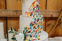 a white wedding cake decorated with colorful dimensional brushstrokes and topped with fresh blooms and greenery