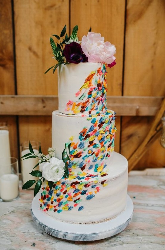 a white wedding cake decorated with colorful dimensional brushstrokes and topped with fresh blooms and greenery
