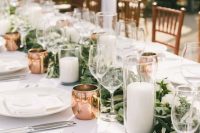 an exquisite wedding tablescape with a greenery and white flower runner, pillar candles, copper mugs, white porcelain and linens