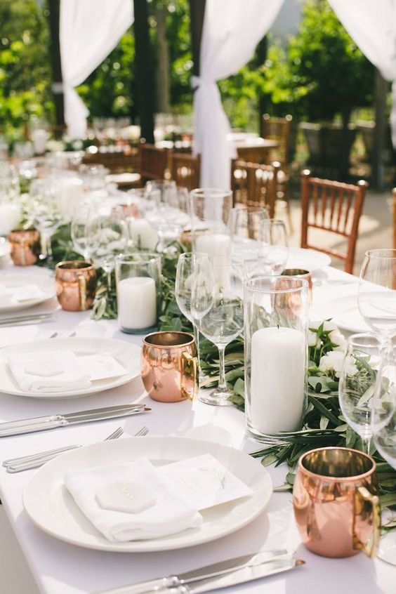 an exquisite wedding tablescape with a greenery and white flower runner, pillar candles, copper mugs, white porcelain and linens
