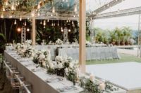 an outdoor spring wedding reception with greenery and bulbs on the beams, white blooms and candles and greenery runners