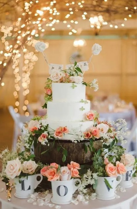 a white buttercream wedding cake decorated with neutral and peachy blooms, greenery, a banner cake topper is a lovely idea for a summer wedding