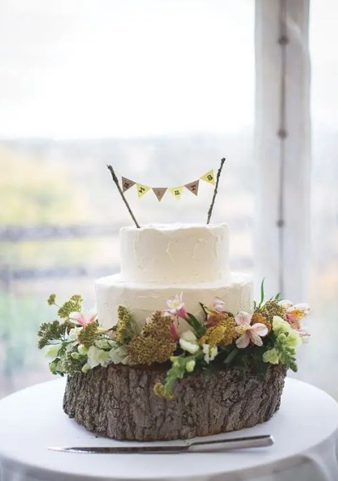 a white textural buttercream wedding cake topped with a banner, on a wood slice with greenery and blooms