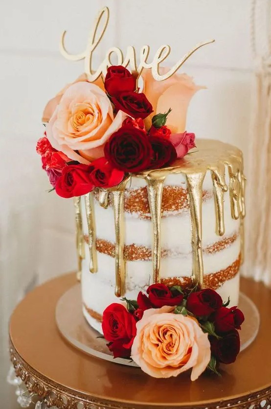 a small naked wedding cake with gold drip, pink, red and burgundy blooms, a gold calligraphy topper is utterly romantic