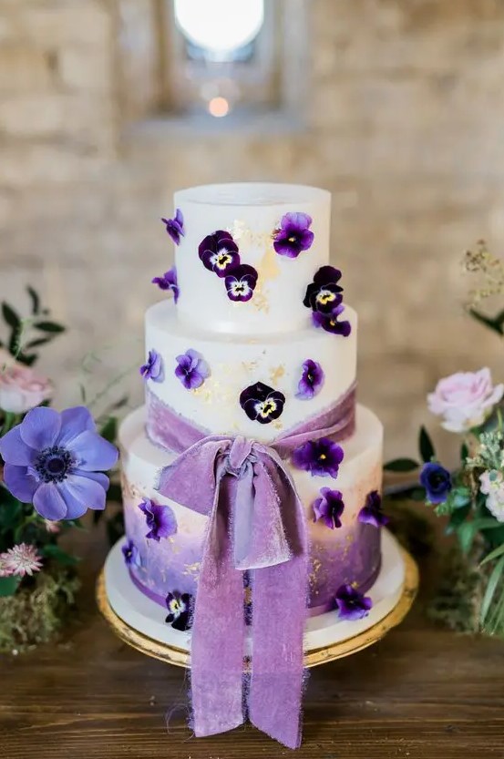 a beautiful ombre white to purple wedding cake decorated with gold leaf, with pansies and a purple ribbon bow is amazing
