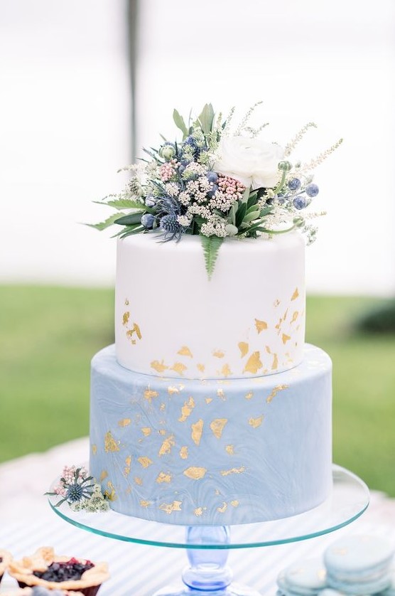 a catchy spring or summer two-tier wedding cake with a white and watercolor blue tier, some greenery, thistles, berries and a bloom on top