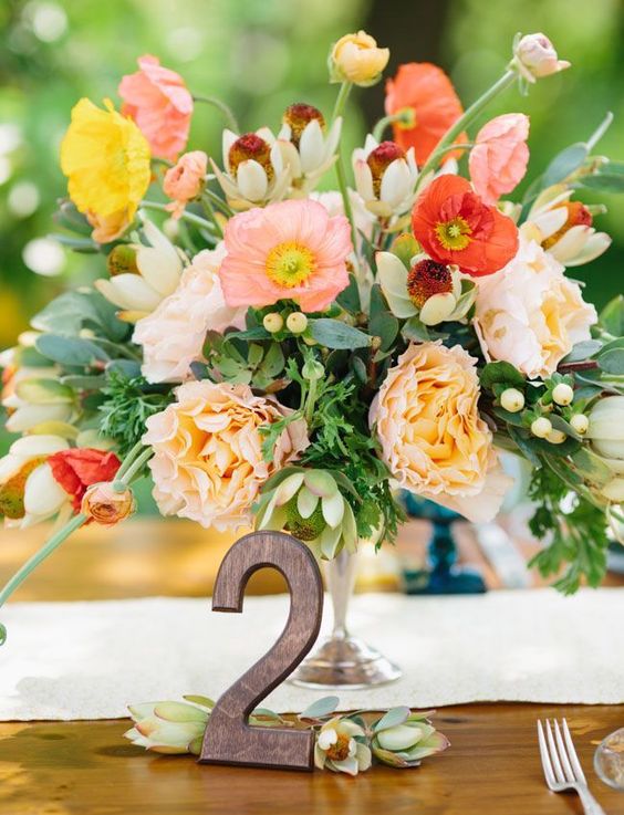 a colorful wedding centerpiece of red, pink, white and peachy blooms and foliage and a wooden table number