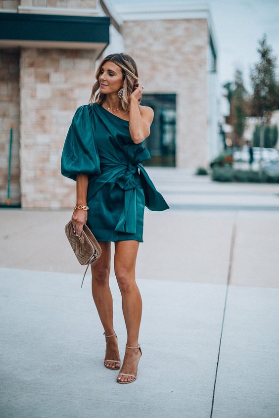 a green one shoulder mini dress with a single puff sleeve, a sash, nude heels and a metallic bag for a summer or fall wedding